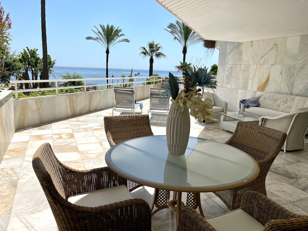 An outdoor terrace is presented with a round glass table surrounded by four wicker chairs. Lush greenery and palm trees are visible in the background, framing views of the sea. A cozy seating area is also included, enhancing the inviting open space.