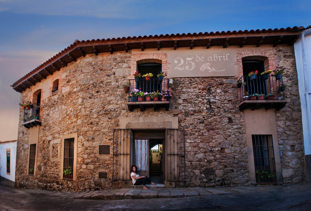 The exterior of an ancient stone house is displayed, featuring a rounded façade with intricate floral decorations on the balconies. The entrance is framed by a large archway, while the building's name, '25 de abril', is engraved above the door. Warm lighting enhances the stone texture.