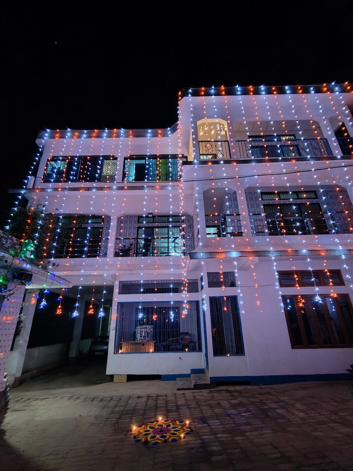 The exterior of a multi-story house is adorned with colorful string lights, illuminating the façade in vibrant hues of blue, white, and orange. The entrance is framed by decorative lanterns, enhancing the festive atmosphere. The night sky serves as a backdrop, highlighting the lit-up structure.