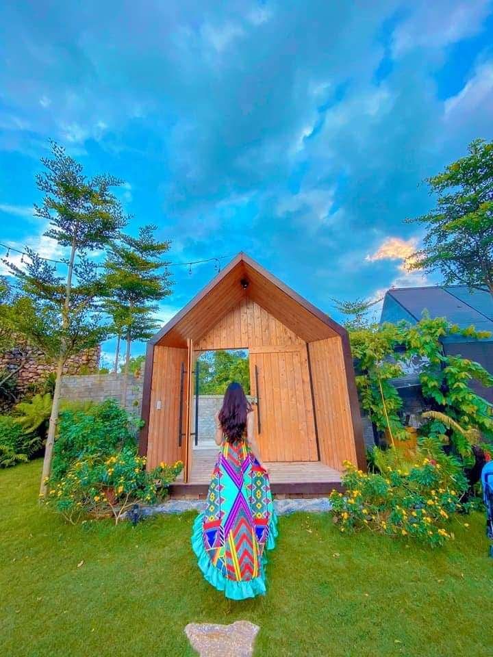 A wooden building with a triangular roof stands amidst lush greenery. The vibrant colors of a patterned garment are seen as a figure approaches the entrance, which is framed by flowering plants and trees under a partly cloudy sky.