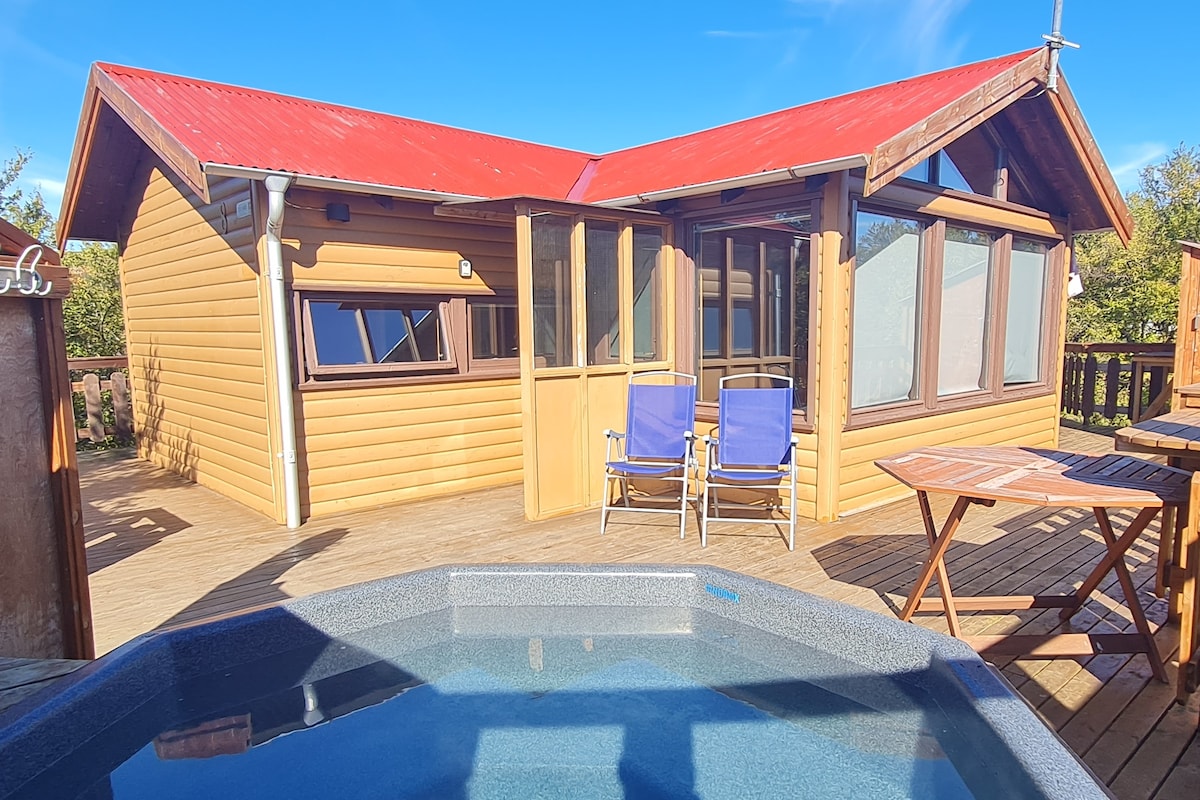 A cabin exterior features a red slanted roof and wooden siding, surrounded by trees. Two blue chairs are positioned on a deck area, alongside a small table. A hot tub is visible in the foreground, offering a relaxing outdoor space.