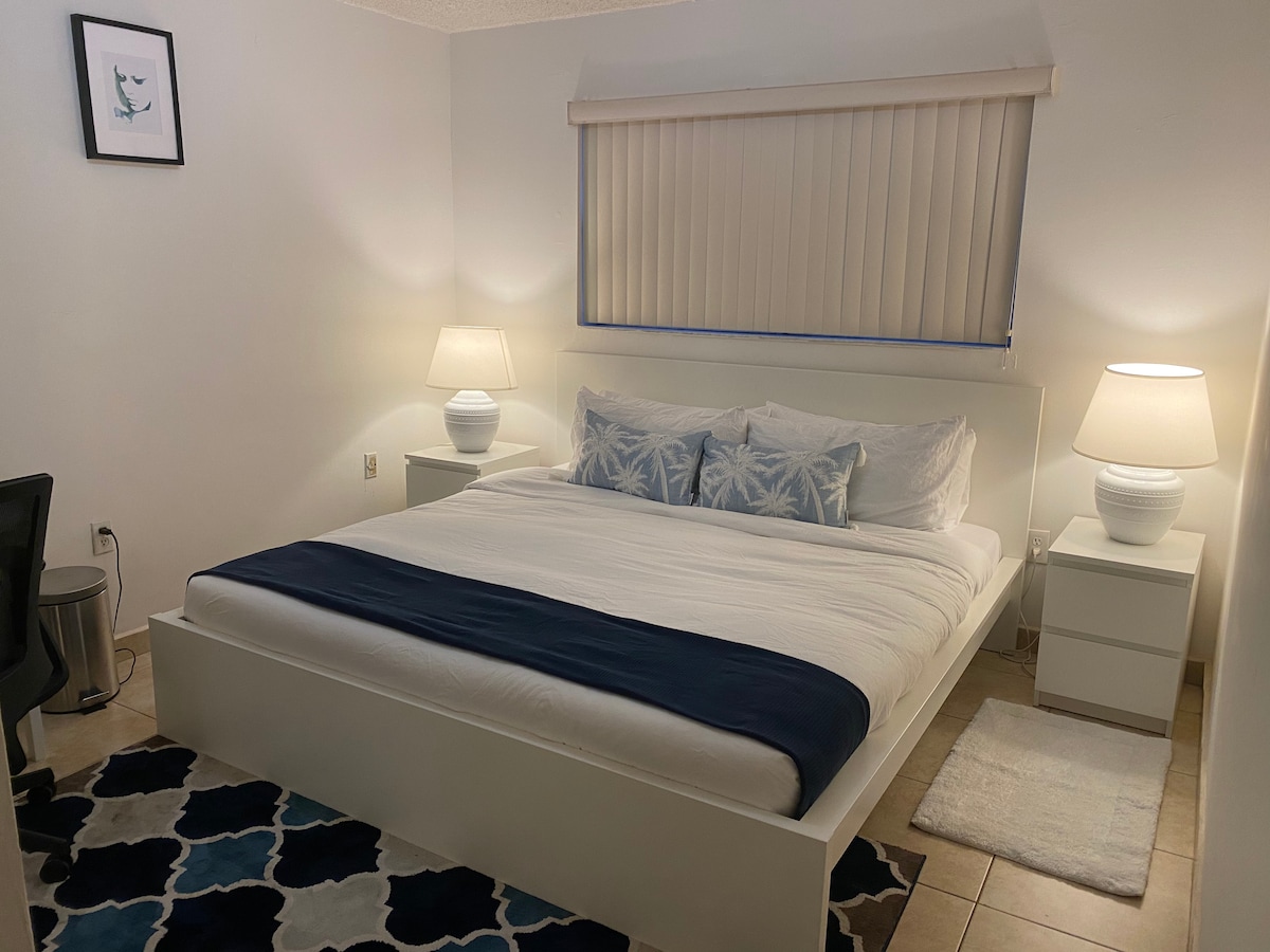 A spacious bedroom is furnished with a large bed featuring white linens and decorative pillows. Two bedside lamps provide soft lighting alongside minimalistic nightstands. A light-colored rug with a pattern lies on the tile floor. A large window with vertical blinds allows natural light to fill the room.