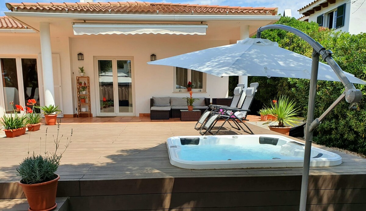An outdoor area is showcased, featuring a hot tub beside a shaded umbrella and two lounge chairs. Potted plants and flowers add a touch of greenery, while covered seating is visible in the background, creating an inviting space for relaxation.