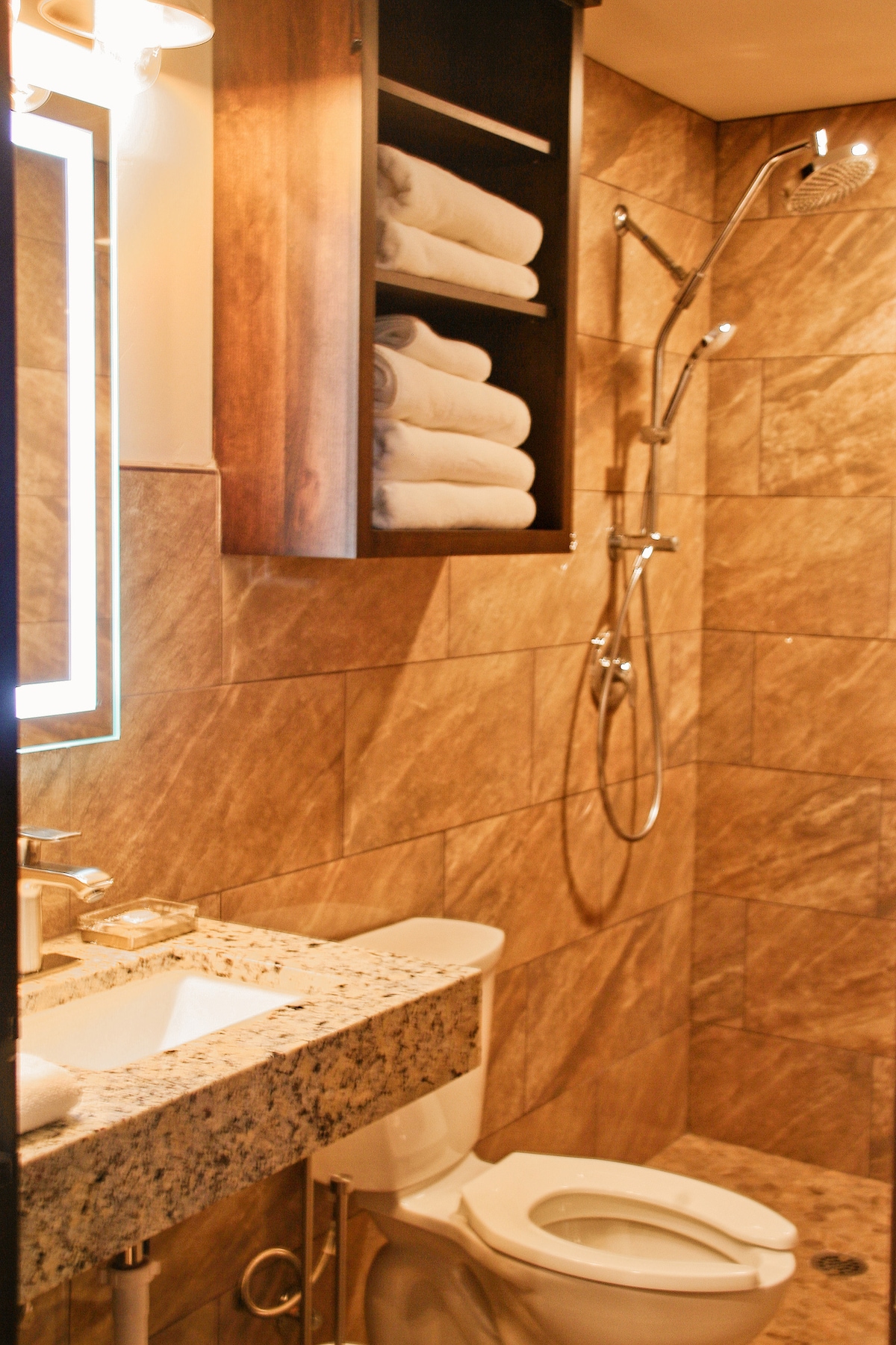 A modern bathroom is presented with warm, earthy-toned tiles covering the walls. The granite countertop holds elegant toiletries, while neatly stacked towels are stored in a cabinet. A shower with a spray head and a toilet are also featured in the space.