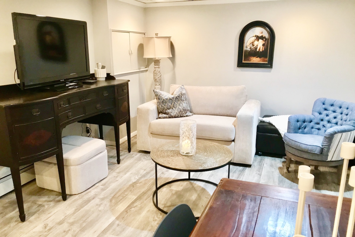 A comfortable living area is presented, featuring a light-colored sofa and a blue accent chair. A round glass coffee table sits at the center, while a television is positioned atop a dark wooden console. Subtle decorative elements are visible, enhancing the welcoming environment.