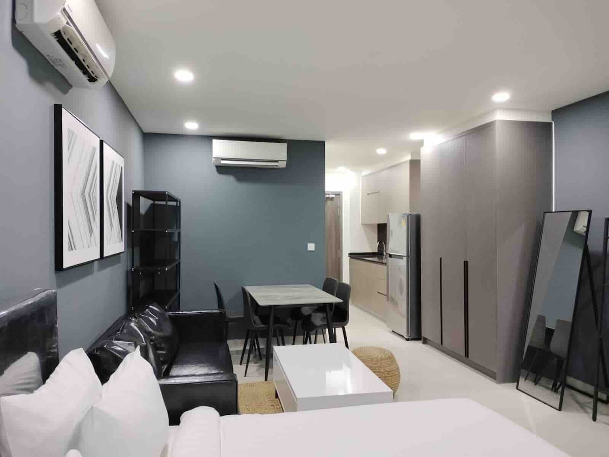 A modern living space features a comfortable sectional sofa and a dining table with four chairs. Light-colored walls and artwork complement the sleek design. The kitchen area is equipped with essential appliances, while a large mirror enhances the room's sense of openness.