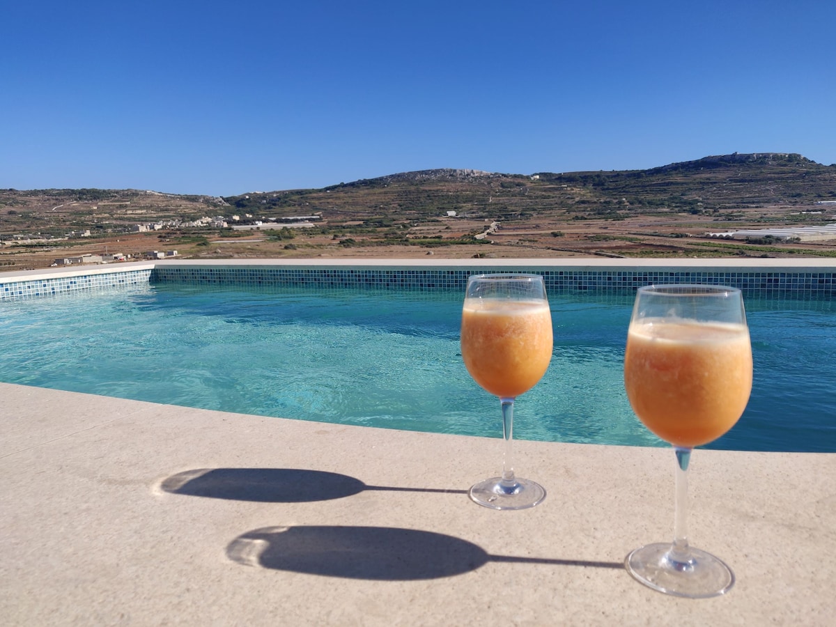 Two glasses filled with a light beverage are positioned on the edge of a private pool. The pool reflects the clear blue sky and offers a view of the surrounding countryside, characterized by gentle hills and distant farmland.