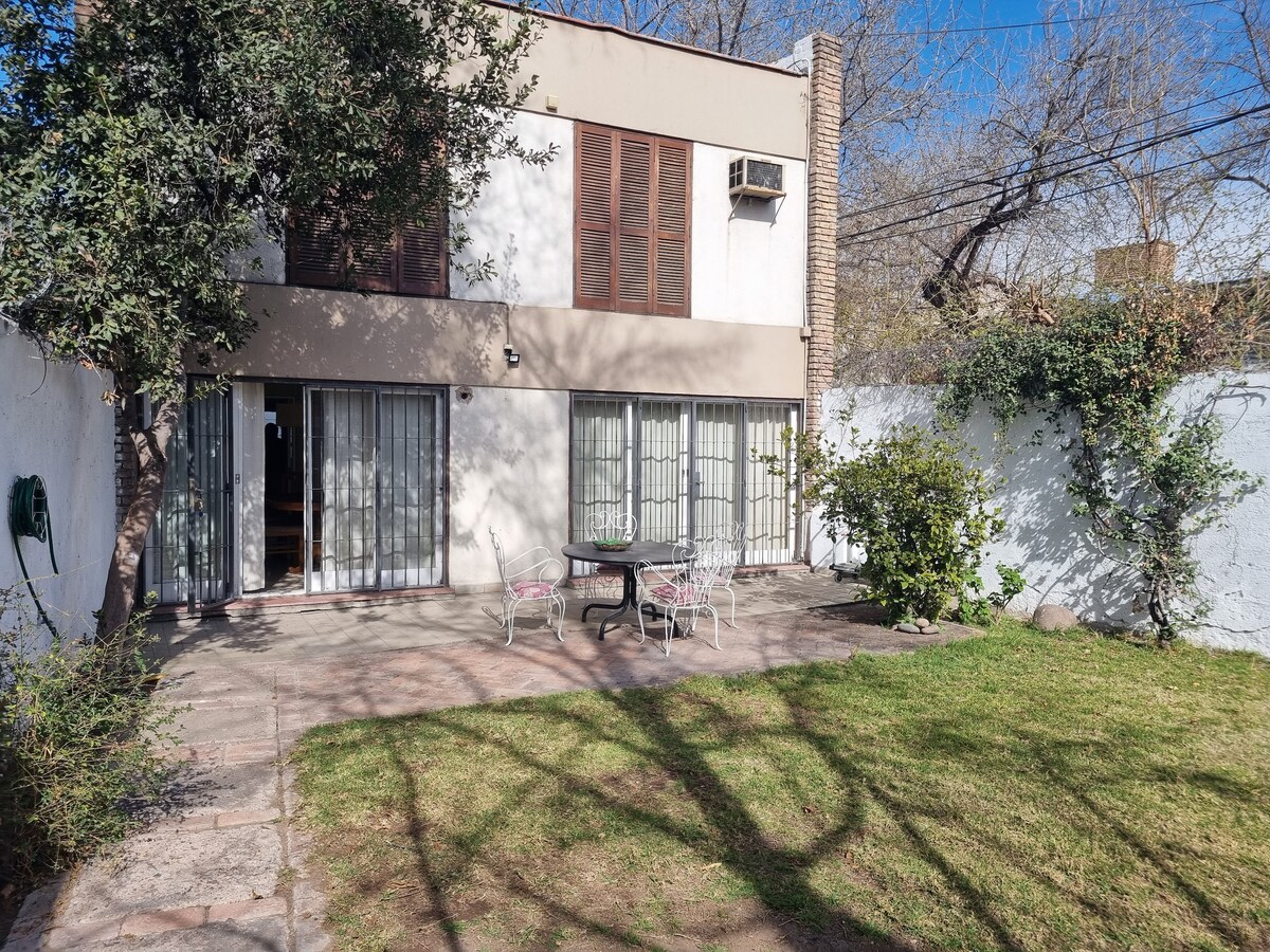 A private outdoor area features a stone patio with a round table and four chairs, surrounded by a well-maintained lawn. Lush greenery and a wall with climbing plants create a serene backdrop. Large glass doors provide access to the interior, inviting natural light.