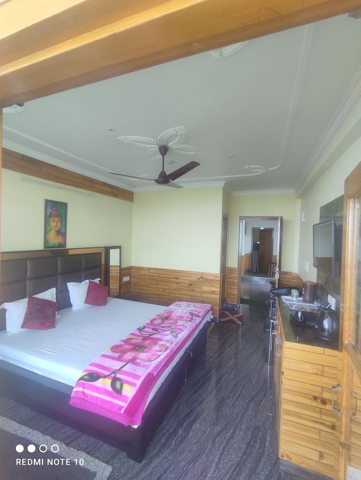 A well-lit bedroom features a spacious bed dressed with a colorful blanket. Wooden accents are visible on the walls and furniture, while a ceiling fan contributes to the room's ventilation. A doorway leads to a separate area, enhancing accessibility.
