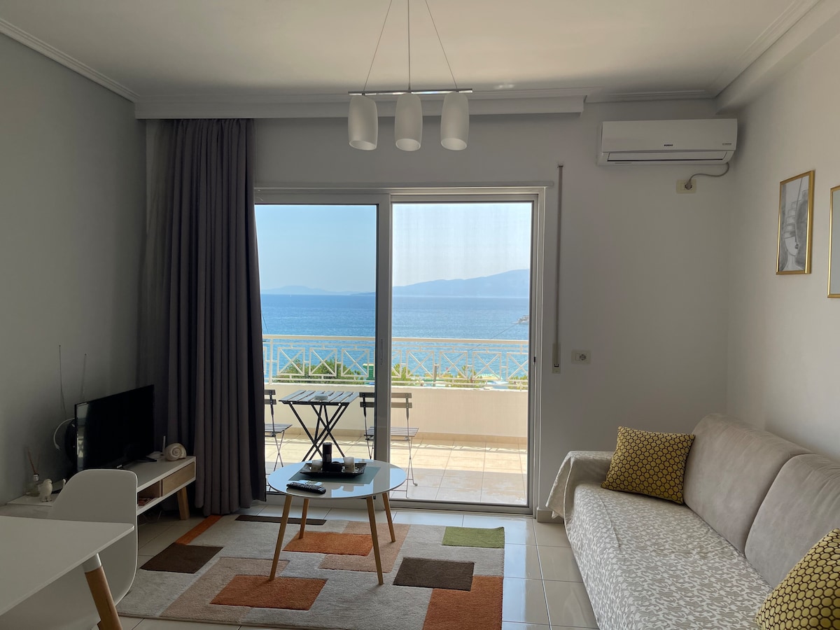 A bright living area features a comfortable sofa and a small table, with a view of the Mediterranean Sea visible through sliding glass doors. Decorative pillows add subtle color, while a small dining table is positioned near the window for enjoying the scenery.