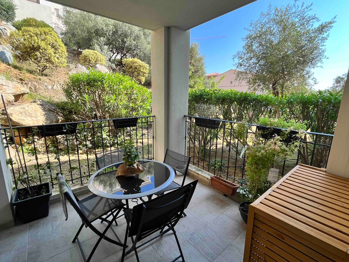 A terrace is visible, featuring a small glass table surrounded by four folding chairs. Potted plants adorn the area, and a wooden storage unit is placed nearby. A garden view is provided, with green foliage outlining the space and a clear sky in the background.