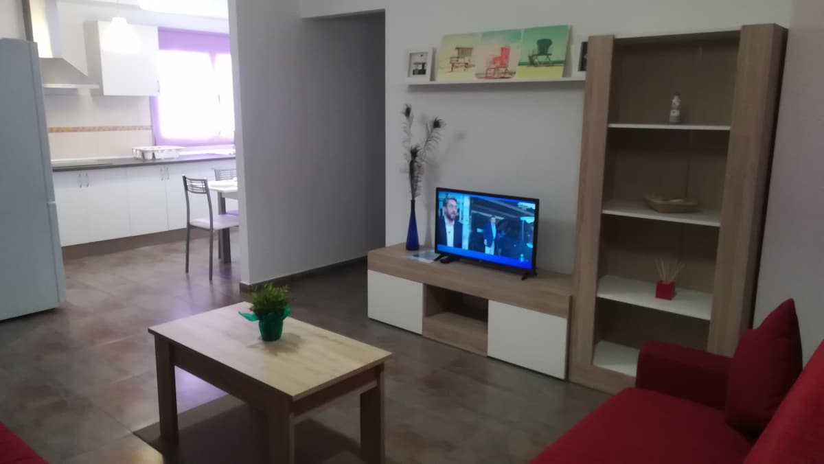 A living area features a red sofa facing a flat-screen TV on a low wooden entertainment unit. A wooden coffee table sits in front, and light streams in through a window, illuminating the space. In the background, a dining table and kitchen are visible.