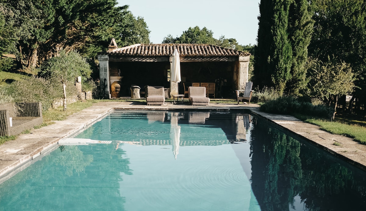 A serene swimming pool is framed by lush greenery and rustic stone pathways. Lounge chairs are positioned on the pool deck, with a shaded area provided by a large umbrella. The backdrop features a charming barn structure, enhancing the peaceful atmosphere.