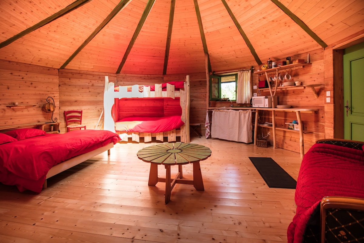 The interior of the wooden yurt features a circular design with a high wooden ceiling. A spacious bed with red bedding is accompanied by two additional beds, also in red. A low round table sits in the center, while a kitchenette area is visible in the background.