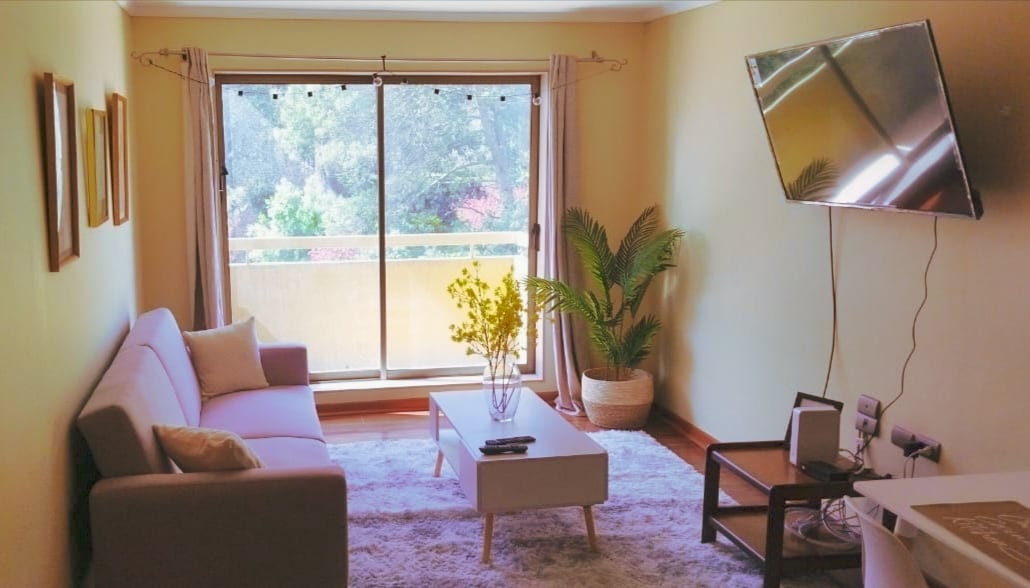 A bright living area features a neutral-colored sofa and a light coffee table centered on a soft rug. Large windows invite natural light, with sheer curtains allowing views of greenery outside. A wall-mounted television is positioned for easy viewing.