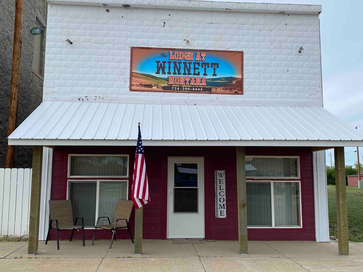 The exterior of the building features a welcoming porch adorned with two chairs and an American flag. A large sign overhead displays the words 'Lodge at Winnett, Montana,' complemented by a simple 'WELCOME' sign at the entrance. The structured façade is primarily white with maroon accents.