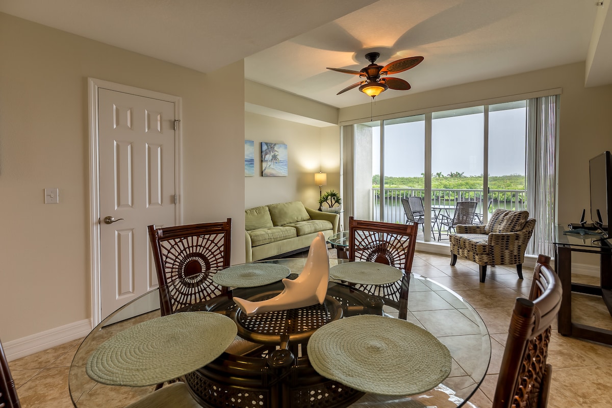 An inviting dining area is set with a round glass table displaying woven placemats and decorative items. Soft natural light fills the space, with views of lush greenery visible through large glass doors that lead to a balcony. A comfortable seating area features a light green sofa.