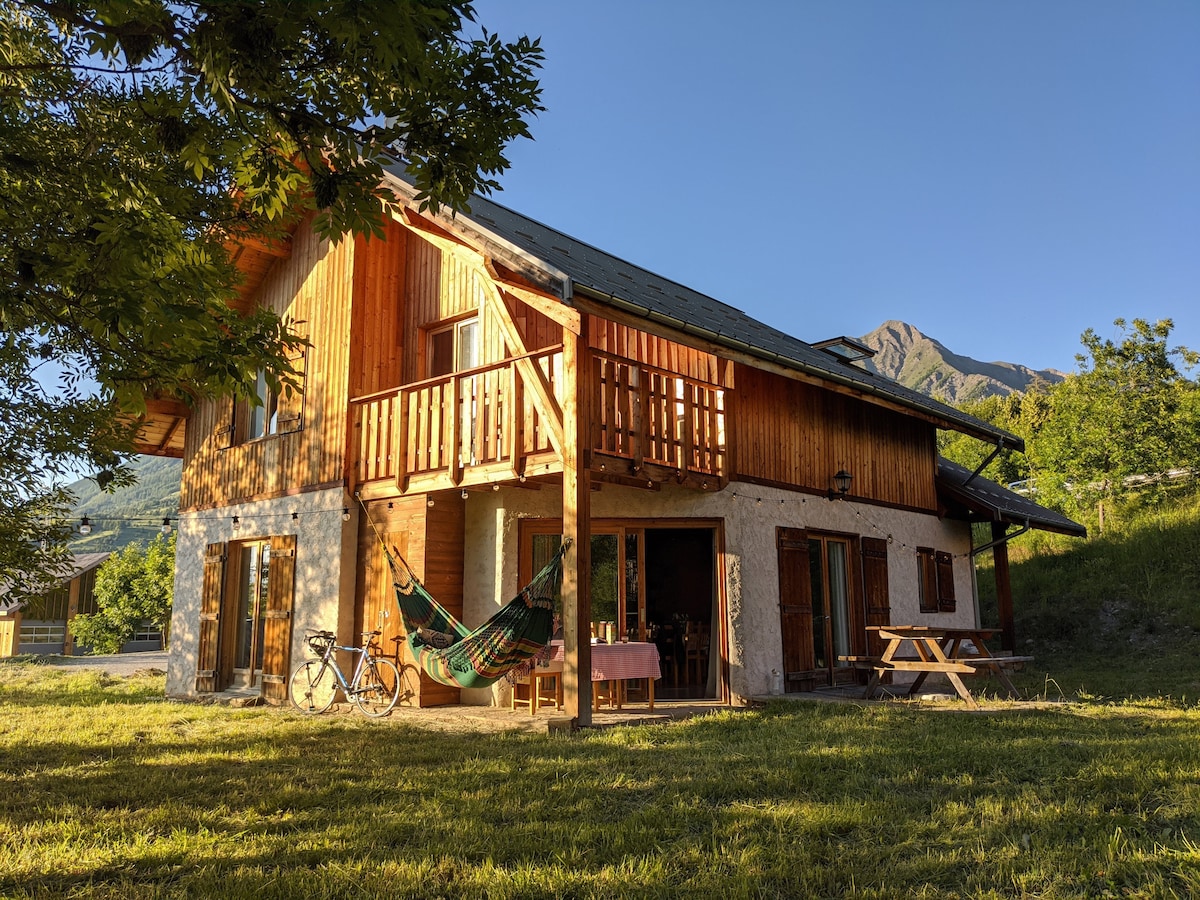 The exterior of a wooden chalet is shown, featuring a spacious balcony and large glass doors. A hammock is hung between two supports, while a picnic table and bicycle are visible on the manicured lawn, with mountains in the background under clear blue skies.
