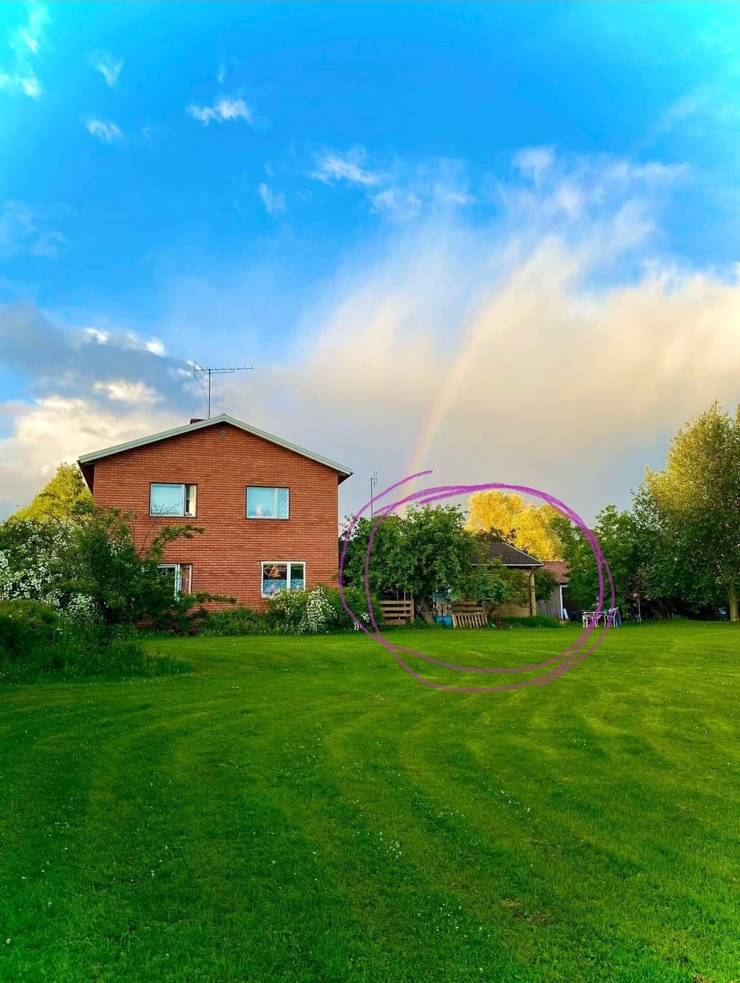 A two-story red brick building is set in a lush green yard under a partly cloudy sky. A faint rainbow is visible above, enhancing the vibrant atmosphere. Trees and a garden area are nearby, featuring seating and a relaxed outdoor space.