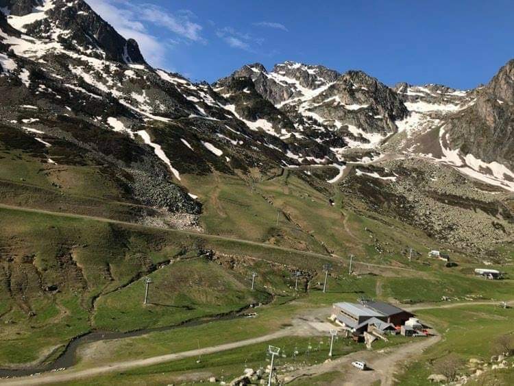 A scenic view of the mountainous landscape features rugged peaks covered in patches of snow and lush green valleys. A small building is visible in the foreground, with a winding road and ski lifts in the distance, conveying the natural beauty of the surrounding area.