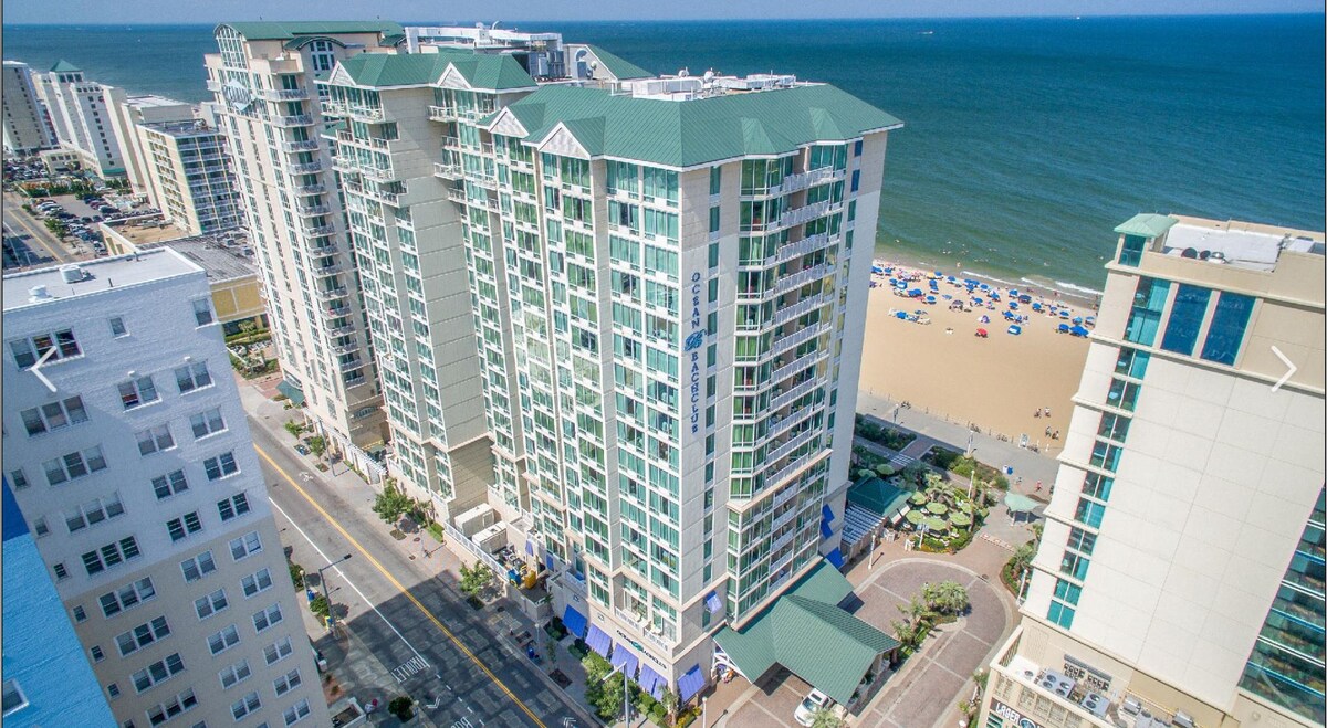 An aerial view captures the beachfront hotel, showcasing its modern architecture against the backdrop of the ocean. The beach area is visible below, dotted with colorful umbrellas. Surrounding buildings highlight the urban landscape of Virginia Beach, with roads leading towards the shoreline.