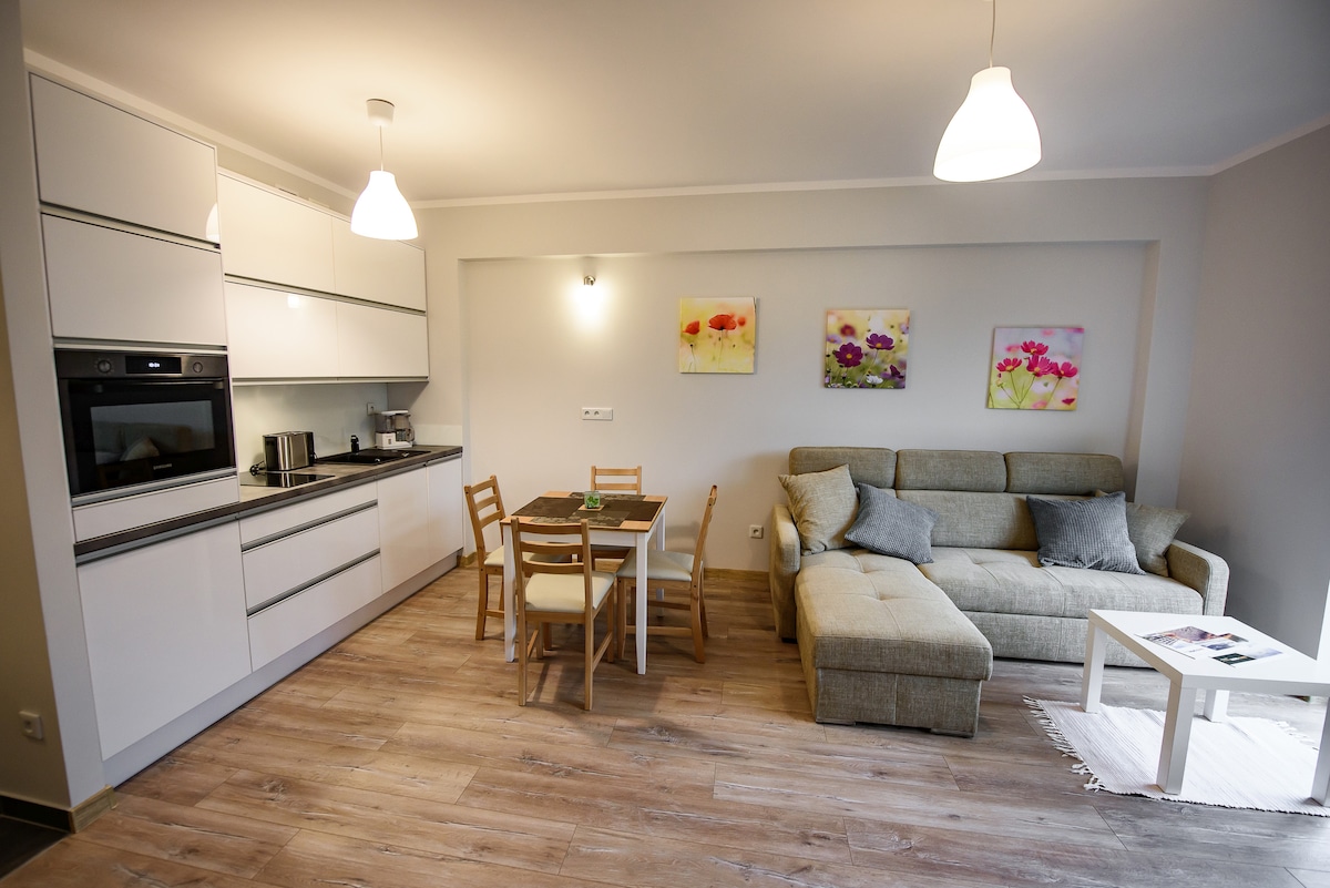 A modern living space features a light-colored kitchenette with built-in appliances, complemented by a cozy dining area with a table and four chairs. A comfortable sofa is situated nearby, adorned with decorative cushions. Floral artwork adds a touch of color to the neutral walls.