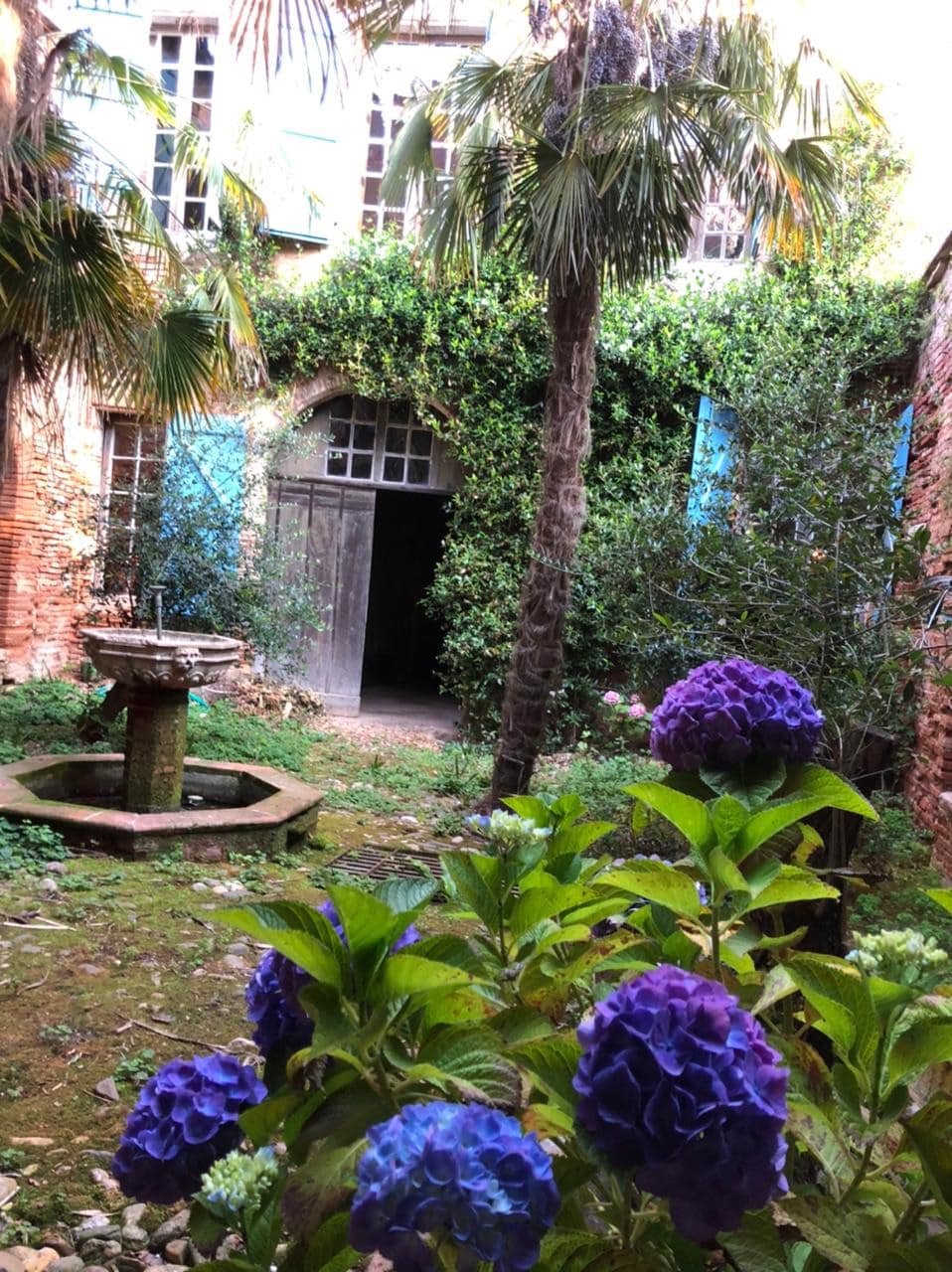 An inviting courtyard is framed by lush greenery and vibrant blue walls. A serene Italian fountain stands at the center, surrounded by flowering plants. The entrance to the apartment can be seen in the background, inviting guests into the historical setting.