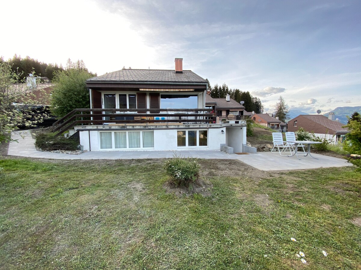 The exterior of the house is shown, featuring a spacious terrace with a railing. Grass surrounds the structure, while trees are visible in the background. The lower level has large windows, allowing natural light to enter. Two white lounge chairs are positioned on the patio.