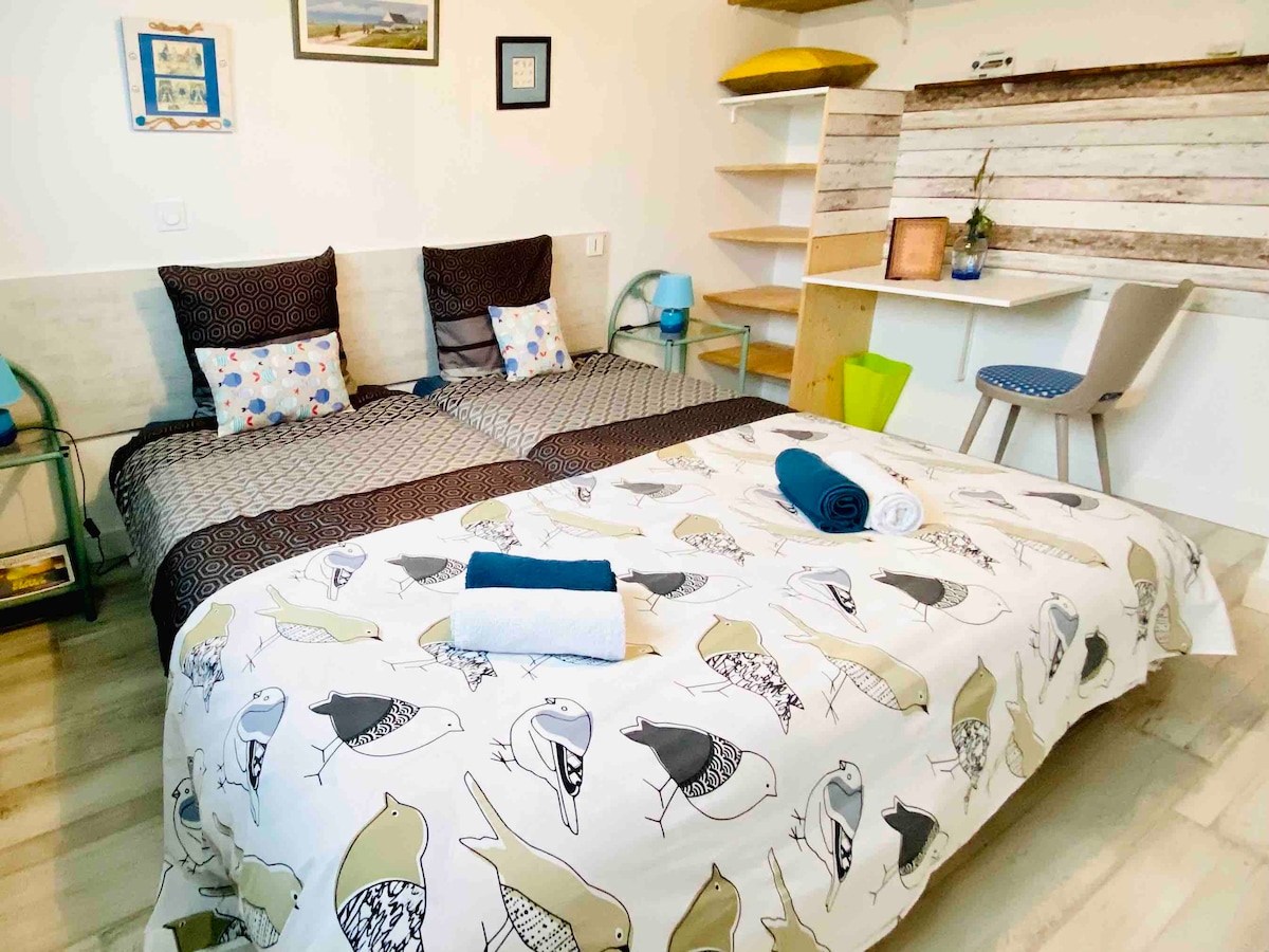 A well-arranged sleeping area features two side-by-side beds dressed in a patterned duvet cover with bird motifs. Neutral-colored walls provide a soft backdrop for decorative elements. A small desk and chair are situated in the corner, complemented by open shelving and a vibrant green accessory.