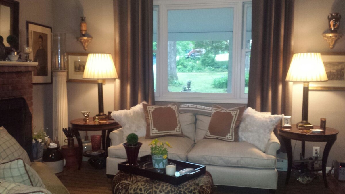 A comfortable living area features a light-colored sofa adorned with decorative pillows. Flanking the sofa are two tables, each hosting a lamp that casts a soft light. A large window offers a view of the greenery outside, enhancing the cozy ambiance.