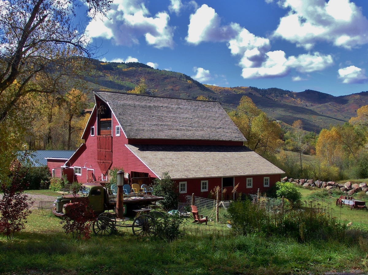 The historic red barn is framed by a scenic landscape featuring rolling hills and autumn foliage. A rustic wagon sits in the foreground, surrounded by greenery. The vibrant blue sky is dotted with fluffy clouds, emphasizing the peaceful country setting.