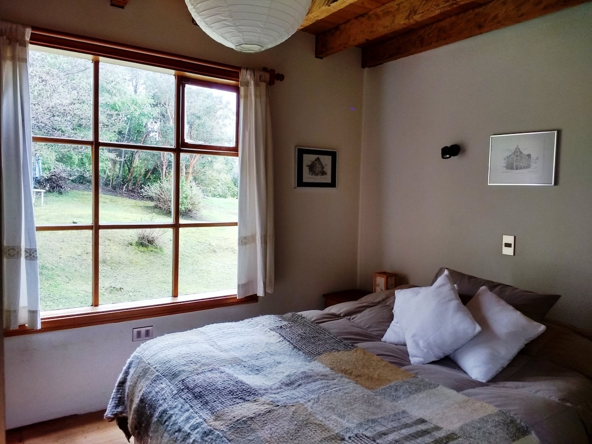 A cozy bedroom is presented with a spacious bed layered in soft blankets and cushions. Natural light filters through a large window, revealing views of greenery outside. Simple decor includes framed artwork on the walls and a bedside table with a lamp.