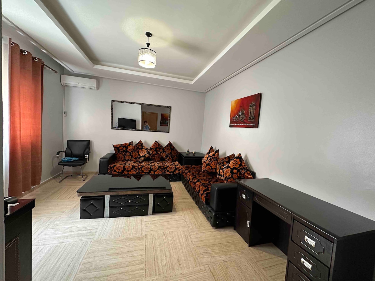A comfortable living area is furnished with a dark sectional sofa adorned with patterned cushions. A sleek coffee table is positioned in front, reflecting soft overhead lighting. A wall-mounted mirror enhances the space, complemented by a decorative artwork above and curtains framing the window.
