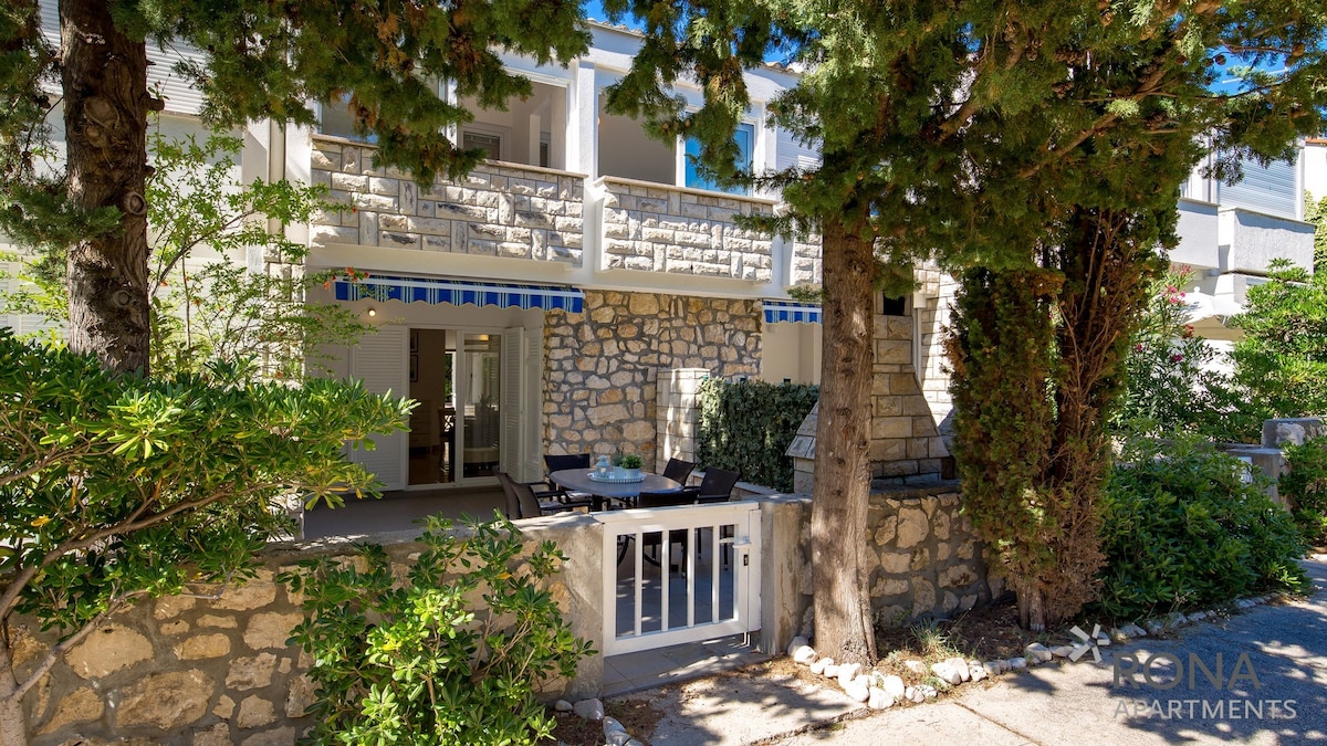 The exterior of the building is framed by lush greenery and stone walls, showcasing a private terrace with outdoor seating. A white gate adds accessibility, while sunlight illuminates the inviting space, creating a serene environment for relaxation.