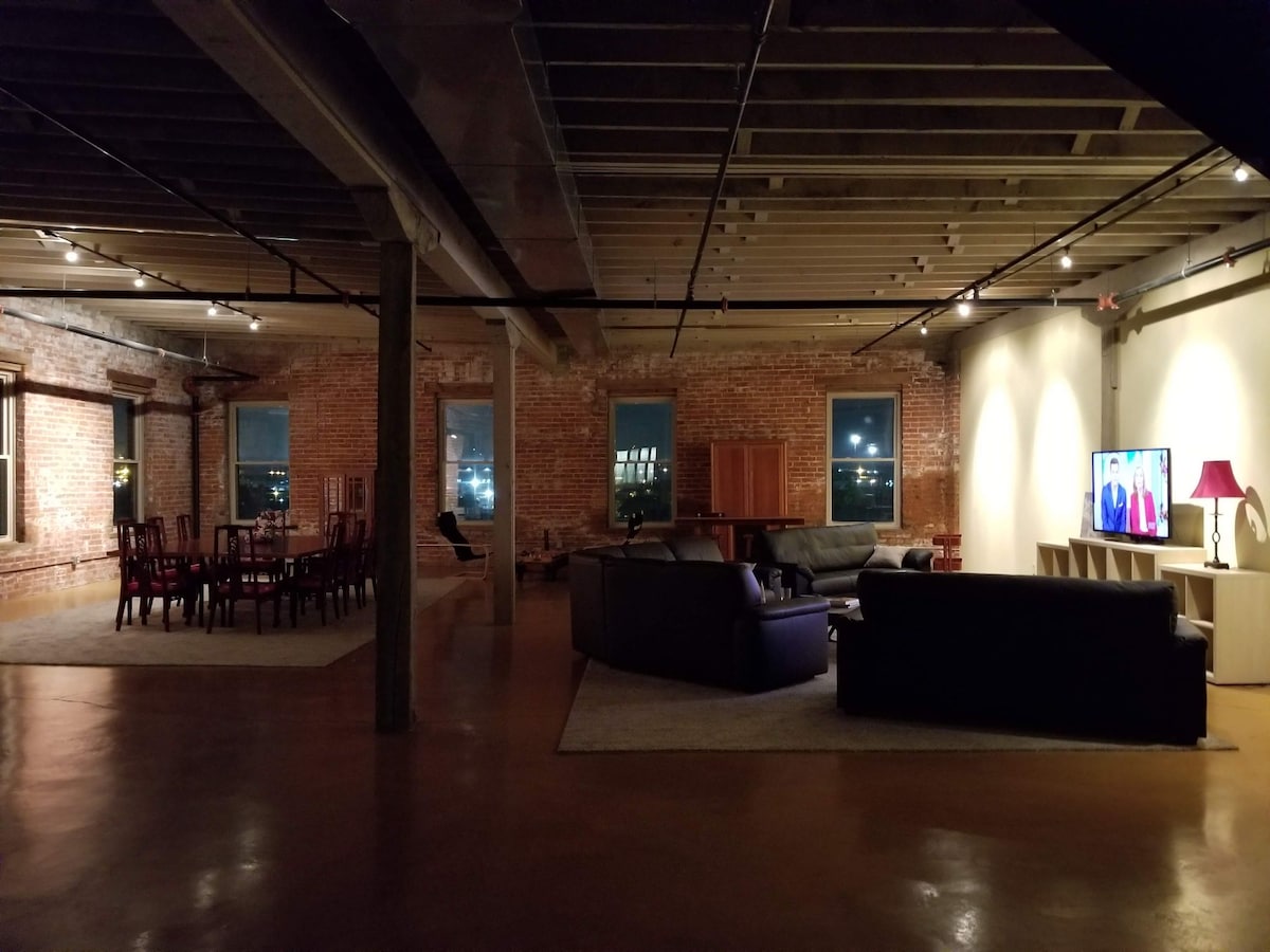 An expansive living area features exposed brick walls and warm-toned lighting. A variety of seating options, including plush sofas, surround a low coffee table. A large dining table accommodates multiple chairs, while a TV is positioned against one wall, creating a comfortable social space.