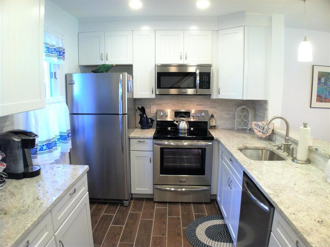 The updated kitchen features stainless steel appliances, including a refrigerator and microwave. White cabinets offer storage, while granite countertops provide ample workspace. A sink is positioned beneath a window, with decorative items adding a touch of style to the light-filled area.