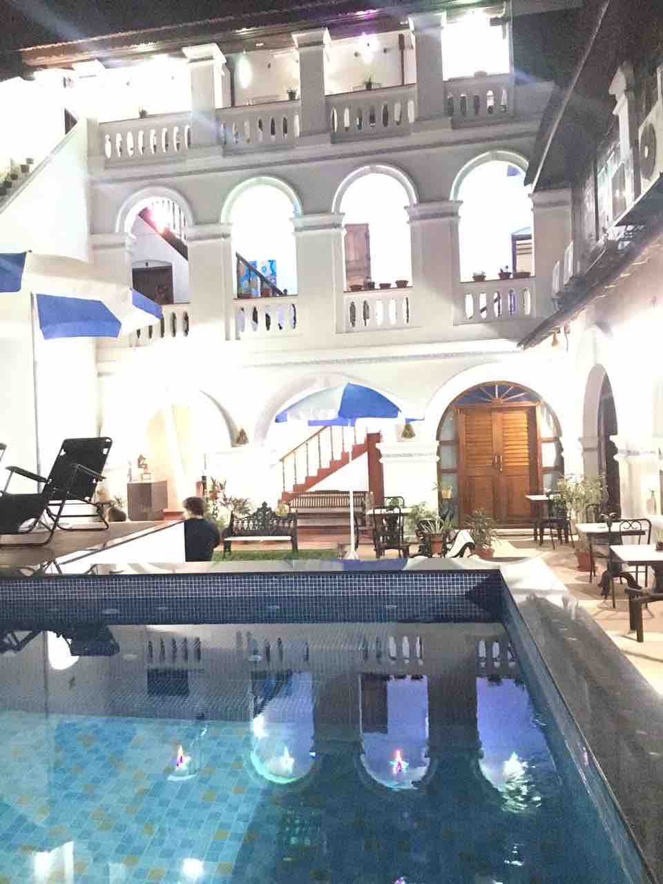 The scene features a beautifully illuminated pool reflecting the light from surrounding architectural elements. Elegant arches frame a staircase leading to the upper levels, while potted plants enhance the courtyard’s ambiance. The tranquil environment invites relaxation and is designed for guest enjoyment.