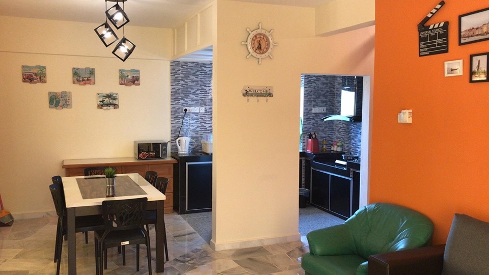 An open living area is highlighted by a dining table with six chairs and a bright orange feature wall. A kitchen area, equipped with a microwave and kettle, is visible in the background, complemented by decorative wall art and a cozy seating option.
