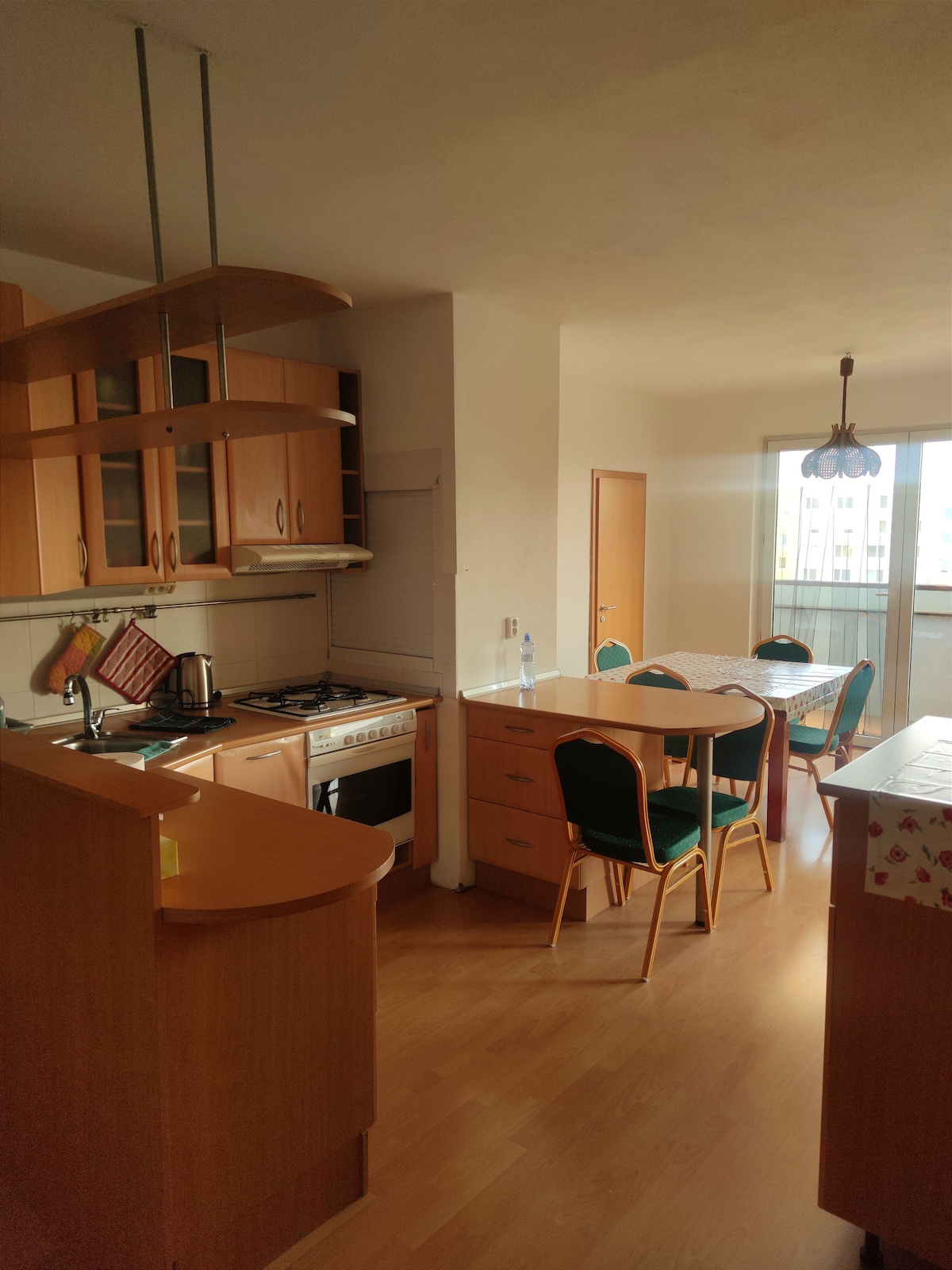 A functional kitchen area is equipped with wooden cabinetry and essential appliances, including a gas stove. A dining table with green chairs is positioned nearby, illuminated by natural light through adjacent windows. The open layout suggests a welcoming space for shared meals.