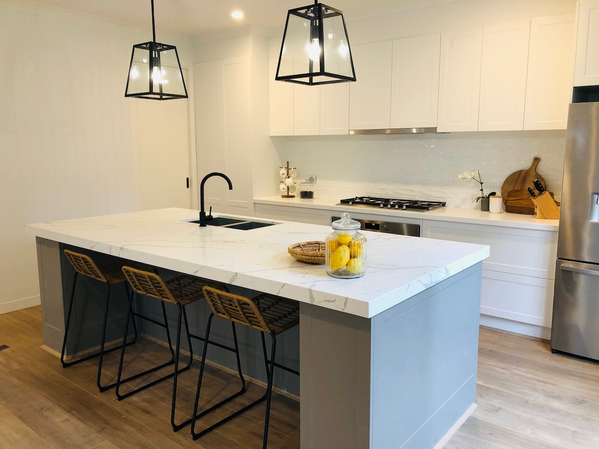 A modern kitchen features a large island with a marbled countertop and three wire-framed stools. Stylish pendant lights hang above, illuminating the space. Sleek cabinetry and stainless-steel appliances complement the minimalist design, creating a functional and inviting cooking area.