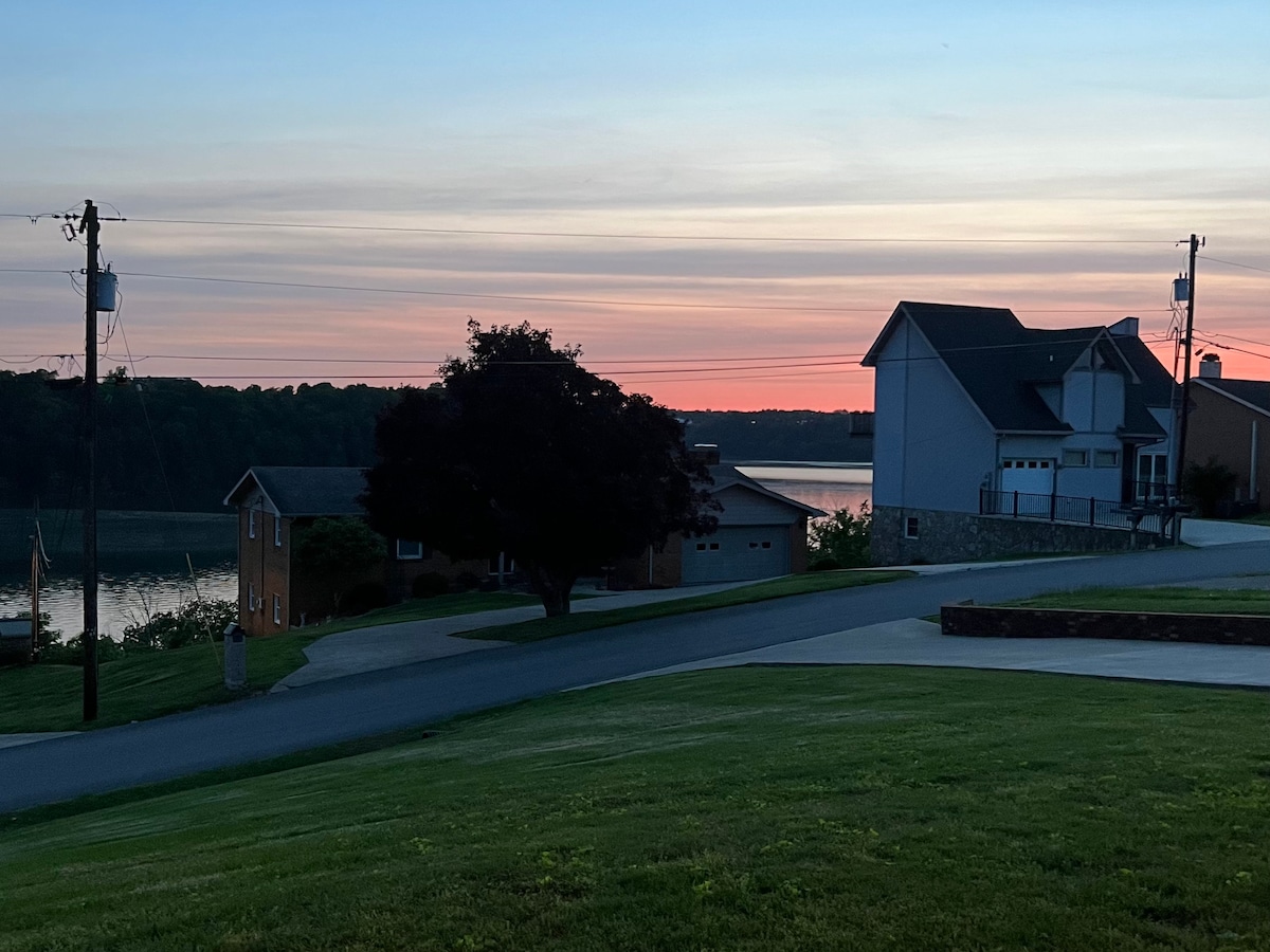 A serene view captures the lake reflecting warm hues of a sunset, framed by lush green grass and a winding driveway. Nearby homes are visible, set against a backdrop of trees and the tranquil waters of Cumberland Lake.