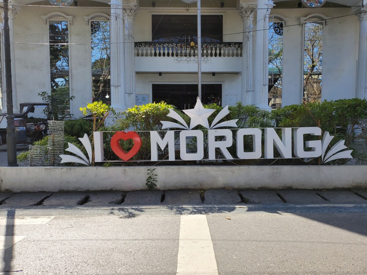 An 'I LOVE MORONG' sign is prominently displayed, featuring bold white letters with a red heart symbol. The sign is set against a backdrop of manicured greenery and a modern building that adds to the local charm.