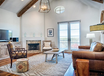 A well-lit living area features a comfortable leather sofa and a stylish wooden chair. A coffee table centers the space, complemented by a patterned area rug. A modern fireplace and a large television are visible, with double doors leading to a scenic balcony.