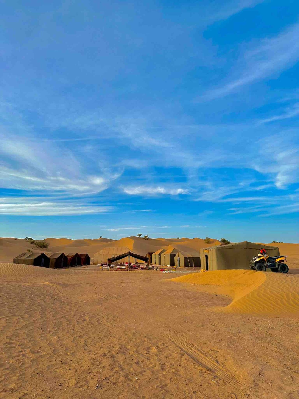 A series of tents is set against the expansive dunes of the Moroccan desert, under a vibrant blue sky with wispy clouds. The scene includes a shaded resting area with seating, and the soft, golden sand is visible in the foreground.