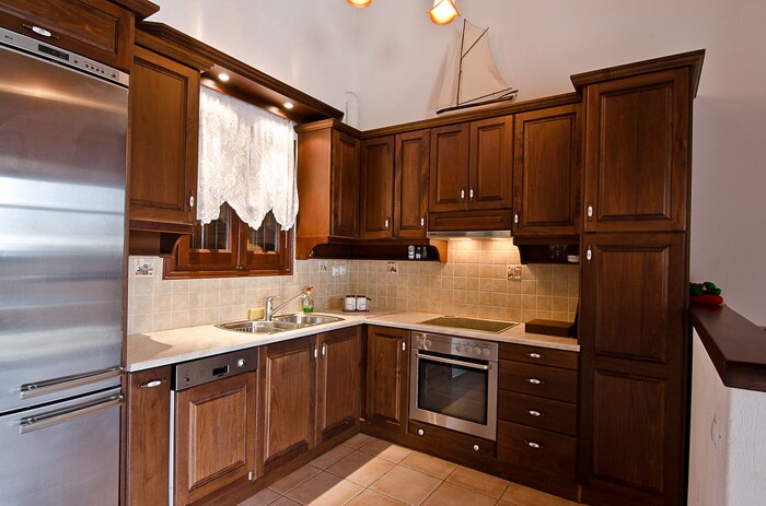 A well-equipped kitchen features rich wooden cabinetry and modern appliances, including an oven and dishwasher. Natural light streams in through a window adorned with lace curtains. The space is complemented by neutral-toned tiles on the floor, providing a warm and functional cooking area.