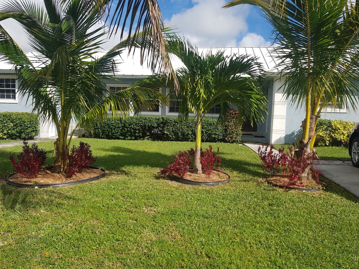 Lush greenery surrounds a well-maintained front yard, featuring several palm trees and colorful landscaping plants. Soft grass covers the ground, inviting relaxation. A clear blue sky with scattered clouds serves as a backdrop for the inviting entrance of the property.