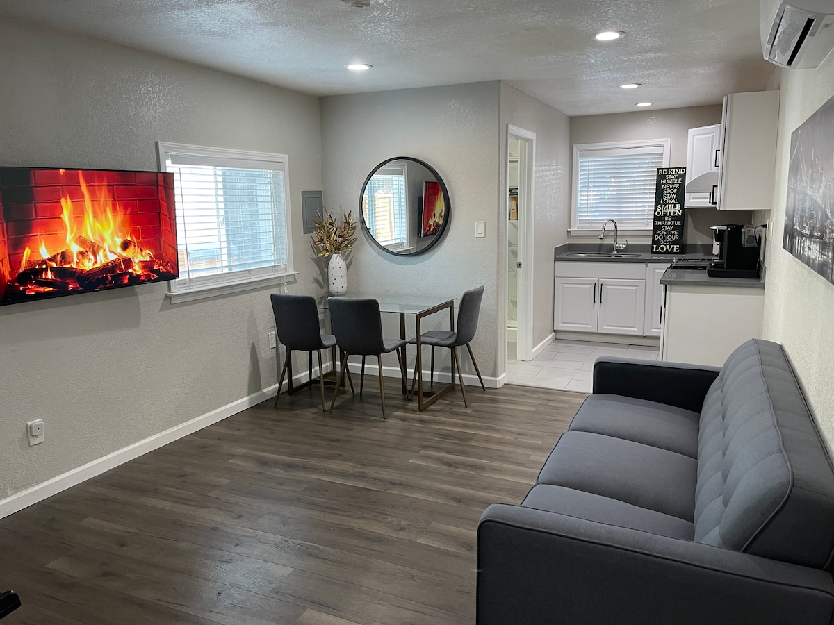 An inviting living space features a comfortable sofa facing a mounted flat-screen TV displaying a virtual fireplace. A round mirror reflects light from the windows. A dining table for four is positioned adjacent to the kitchen, which includes modern appliances and countertops.