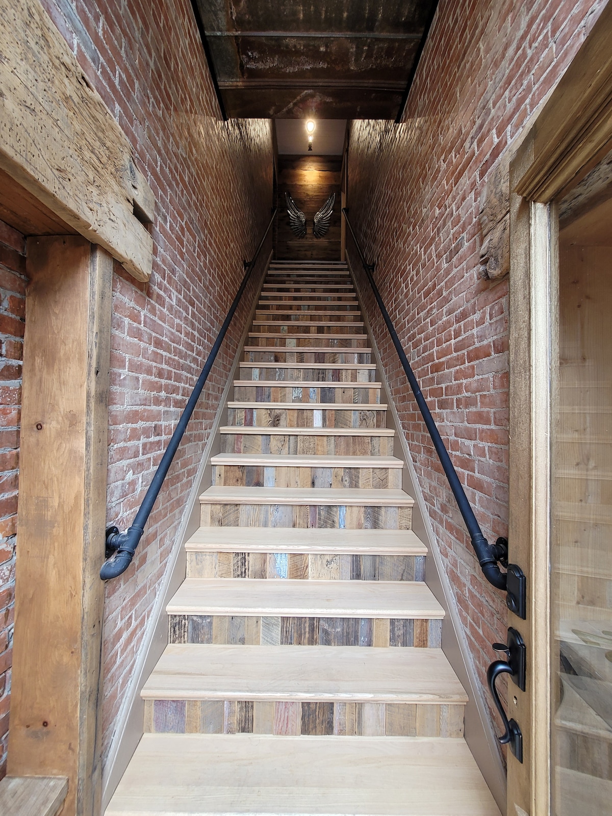 A staircase is featured, leading upward with wooden steps and black metal railings. Exposed brick walls provide a rustic backdrop, while soft lighting illuminates the path. The space above includes wooden beams, adding to the historic charm.