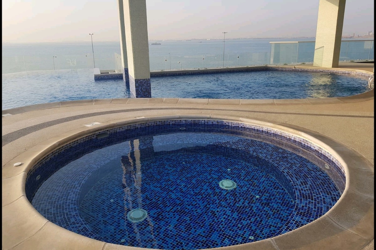 A jacuzzi is surrounded by a smooth concrete deck, displaying vibrant blue tiles that contrast with the water's reflection. In the background, an infinity pool merges with the horizon, offering views of the distant sea and a clear sky.