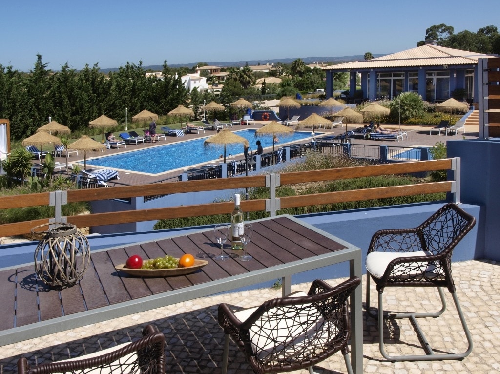 A private terrace features a dining table and chairs, showcasing a view of the resort's swimming pool and sun loungers under straw umbrellas. Lush greenery surrounds the area, and a clear blue sky extends in the background.